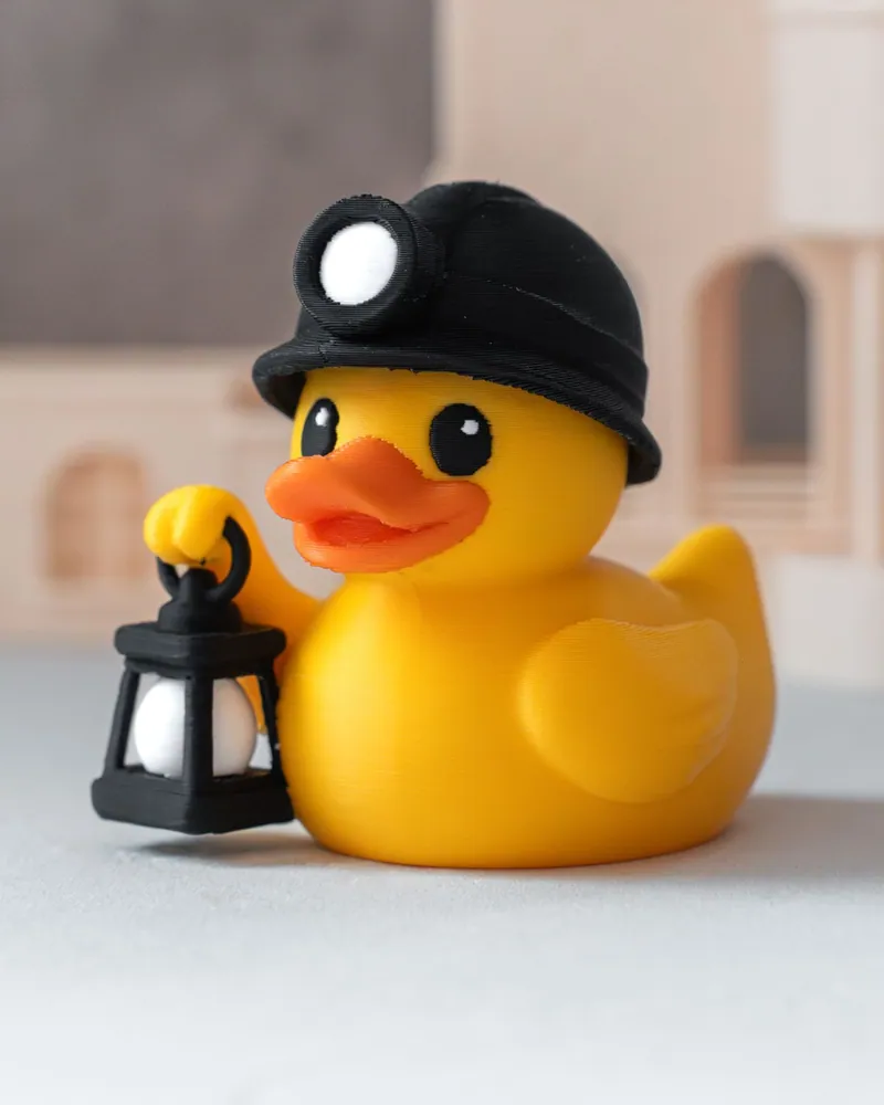 Gele Koempel-eend met zwarte mijnwerkershelm en lamp, met Mijnschacht Nulland op de achtergrond
